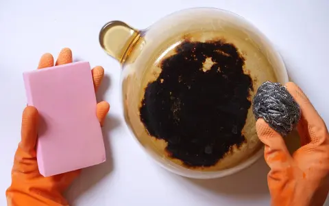 Manos con guantes naranjas sostienen una esponja rosa y un estropajo metálico, listos para aprender como quitar el quemado de una olla.