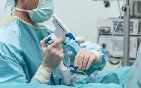 Lekarz w masce i rękawiczkach przygotowuje się do zabiegu, trzymając urządzenie do biopsji. Czy biopsja prostaty może uaktywnić raka?
