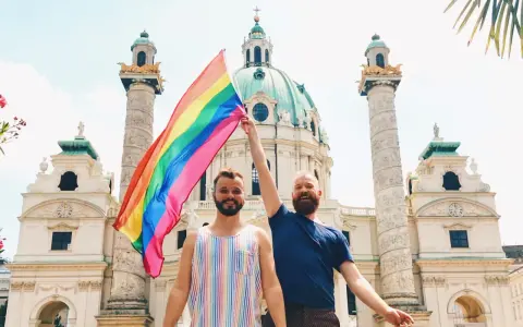 Two men proudly hold a rainbow flag in front of a grand building in Austria, celebrating gay pride.