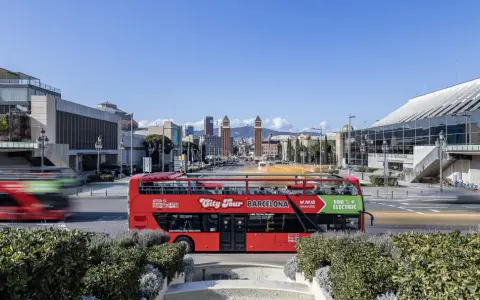 A red double-decker bus, the best hop on hop off Barcelona, drives past modern architecture under a clear blue sky.