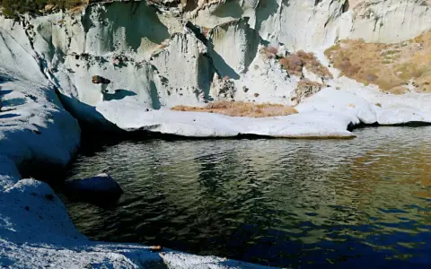 Scogliere bianche erose dal mare, con vegetazione secca e un'acqua scura che riflette il cielo. Sembra un luogo selvaggio, quasi un "cane malu" che custodisce la costa.