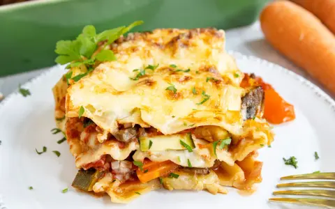 Ein Stück herzhafte vegetarische Lasagne mit geschmolzenem Käse und frischer Petersilie, daneben Karotten.