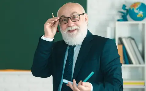 Uśmiechnięty starszy nauczyciel w okularach, z białą brodą, trzymający długopis. Myśli o zasłużonych emeryturach dla nauczycieli.
