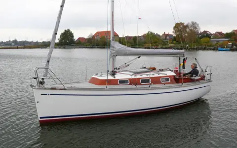 Ein Mann steuert die Segelyacht BIGA 242 auf ruhigem Wasser, im Hintergrund Häuser und Bäume.