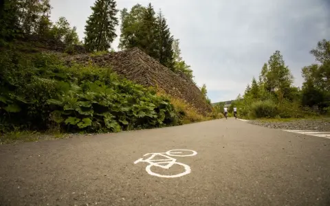 Radweg mit Radsymbol auf Asphalt. Zwei Radfahrer fahren auf einem asphaltierten Weg, der an einer steilen, mit Steinen bedeckten Böschung und üppiger Vegetation vorbeiführt.