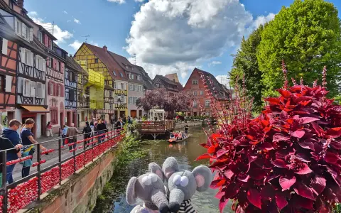 Deux peluches souris posent devant les maisons colorées de Colmar, une destination idéale pour visiter Colmar en 1 jour. Une barque navigue sur le canal.