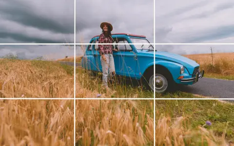Une femme souriante, chapeau de paille, pose près d'une voiture bleue vintage au milieu d'un champ de blé doré. Photos prises sur le vif.