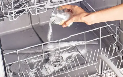 A hand pours white powder from a jar into an open dishwasher, demonstrating how to clean a dishwasher naturally.