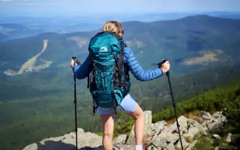 Kobieta z turkusowym plecakiem w góry, z kijkami trekkingowymi, podziwia panoramę.