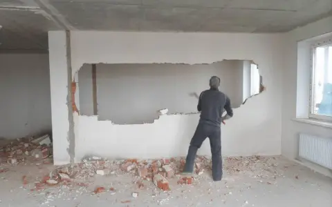 Mężczyzna wyburza młotem duży fragment ściany nośnej, tworząc otwór. Wokół leżą gruzy.