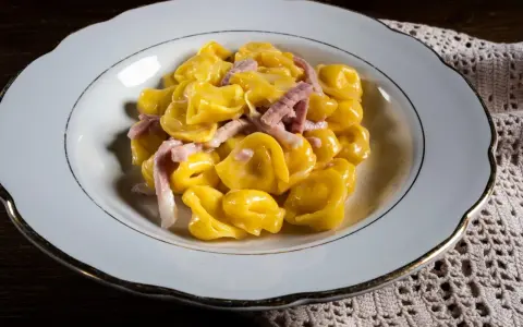 Tortelli ripieni in salsa cremosa con prosciutto, un classico tra i piatti tipici di Parma, serviti in un piatto bianco bordato d'oro.