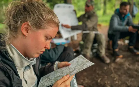 Frau studiert Karte, während Männer im Hintergrund bei einer Ausbildung im Wald sitzen. Solche intensiven, kurzen **ausbildungen die nur 1 jahr dauern** sind ideal für schnelle Karriereschritte.