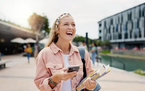 Szczęśliwa studentka z polską maturą, która planuje studia za granicą, śmieje się, trzymając telefon i książki.