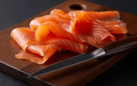 Finas lonchas de salmón ahumado, listas para disfrutar, reposan sobre una tabla de madera junto a un cuchillo.