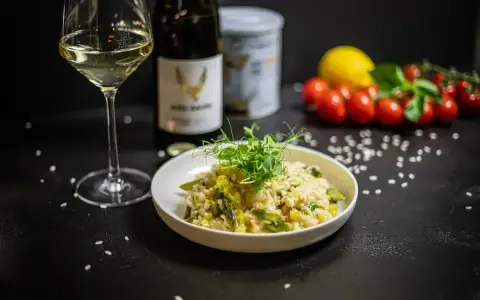 Aksamitne risotto z kurczakiem i szparagami, udekorowane kiełkami. Obok kieliszek białego wina i butelka.