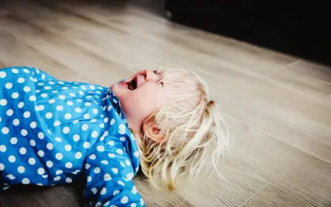 Mała blondynka w niebieskiej sukience w kropki leży na podłodze, krzycząc. To klasyczny bunt dwulatka, gdy świat nie działa tak, jakby chciał.