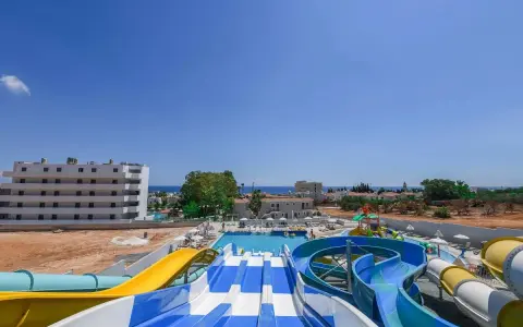 Kolorowe zjeżdżalnie wodne i baseny w Narcissos Waterpark Resort, z hotelami i morzem w tle.