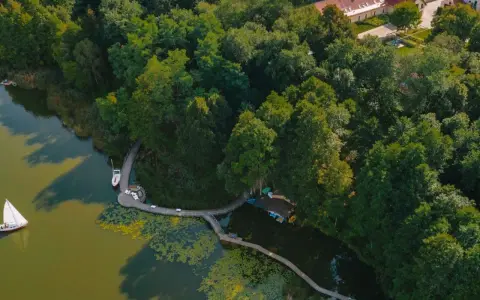 Żaglówka i deska SUP na spokojnym jeziorze ostrowskim, otoczonym bujną zielenią i drewnianą kładką.