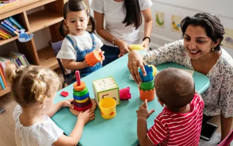 Zwei Frauen und drei Kleinkinder spielen mit bunten Spielsachen. Eine Frau hilft einem Kind, Türme zu stapeln, was eine tolle Methode ist, um Kinder zweisprachig zu erziehen.