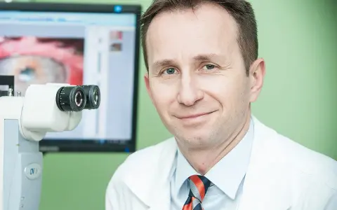 Okulista z uśmiechem przy aparaturze do badań. Na ekranie widok oka, sugerujący diagnostykę przed przeszczepem rogowki.