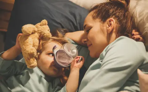 Mama opiekuje się dzieckiem z rotawirusem. Podaje mu wodę z kubeczka, a obok leży pluszowy miś.