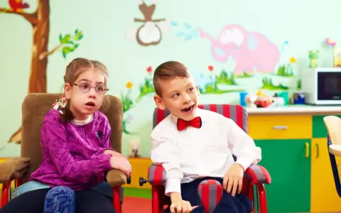 Dwoje dzieci w fotelikach, dziewczynka w okularach i chłopiec w muszce, na tle kolorowej ściany z rysunkami.