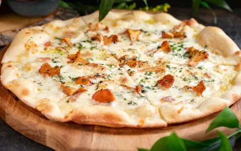 Pyszna pizza z kurkami, polana kremowym sosem i posypana świeżymi ziołami, podana na drewnianej desce.