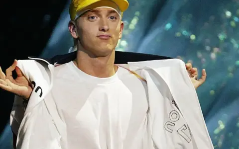Slim Shady w żółtej czapce i białej bluzie, z gestem dłoni.