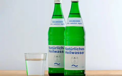 Zwei grüne Flaschen Heilwasser und ein Glas Wasser. Ein Glas Wasser vor dem Schlafengehen trinken kann beim Abnehmen helfen.