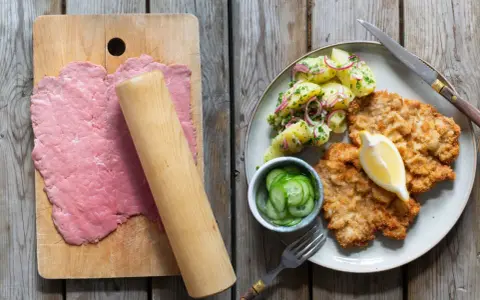 Förberedelser för schnitzel recept: tunt bankat kött på skärbräda, bredvid en tallrik med schnitzel, potatissallad och gurka.