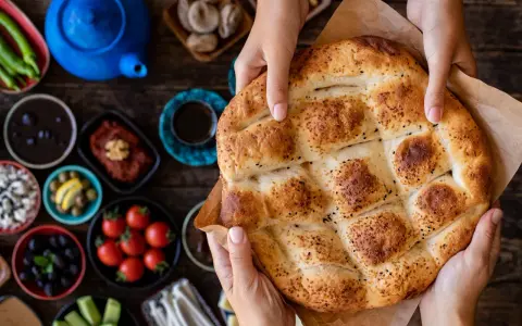 Hände halten ein frisch gebackenes Ramadan Brot, bestreut mit Sesam und Schwarzkümmel. Daneben liegen kleine Schalen mit Oliven, Tomaten und anderen Köstlichkeiten.