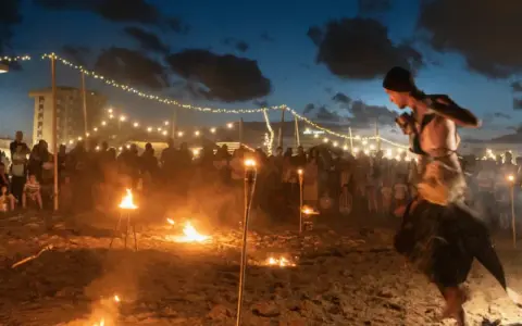 Spettacolo di fuoco sulla spiaggia di Rimini, estate. Artista si esibisce tra torce e fuochi d'artificio, con pubblico e luci in lontananza.