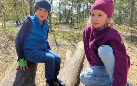 Två barn sitter på stockar i skogen. Båda bär en mjuk merinoull mössa barn, perfekt för utomhuslek.