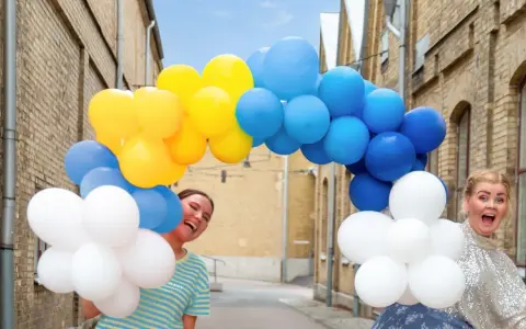 Två glada personer gömmer sig bakom en båge av gula, blå och vita ballonger. Perfekt för studentmottagning lekar!