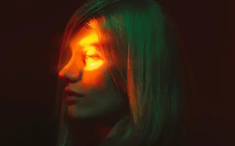 Profilo di donna con luce arancione sull'occhio, evocando un'emicrania oculare. Capelli illuminati da luci verdi e rosse.