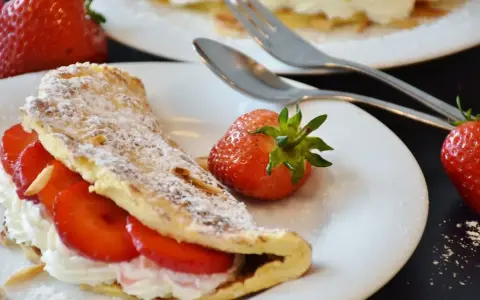 Puszysty omlet z białek, nadziany kremem i truskawkami, posypany cukrem pudrem. Obok leżą świeże owoce i sztućce.