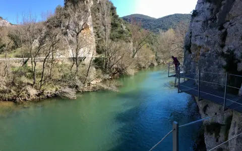 Persona camina por pasarela metálica junto a río turquesa, explorando las rutas de Burgos. Paisaje rocoso y vegetación de ribera.