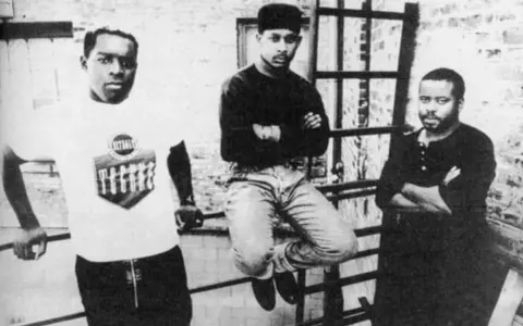 Trzech mężczyzn, pionierzy detroit techno, pozuje na tle ceglanej ściany.
