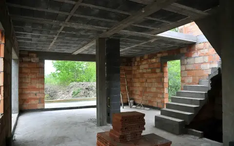 Wnętrze budowy z widocznym stropem gęstożebrowym, schodami, ceglanymi ścianami i otwartymi drzwiami na zewnątrz.