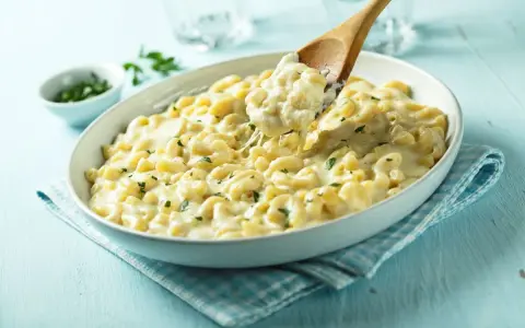 Pyszne, kremowe mac n cheese z ziołami, podane w białej misce. Drewniana łyżka nabiera porcję ciągnącego się sera.