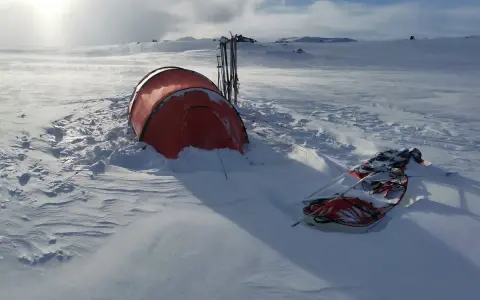 Ein rotes Wintercamping Zelt steht im Schnee, daneben Skier und ein Schlitten. Die Landschaft ist weit und verschneit.