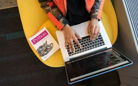 Osoba w pomarańczowej kurtce pisze na laptopie obok książki o Pythonie, siedząc na żółtym krześle.