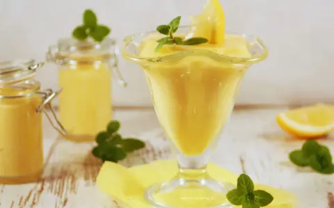 Pyszny lemon curd przepis w pucharku, ozdobiony cytryną i miętą. Obok słoiczki z kremem.