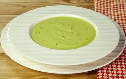 Una ciotola di cremosa crema di zucchine, condita con pepe nero, servita su piatti bianchi su un tavolo di legno.