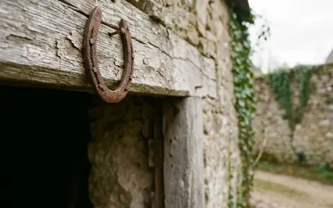 Un vieux fer à cheval pointes en bas, rouillé, est cloué sur une poutre en bois patiné, devant une bâtisse en pierre recouverte de lierre.