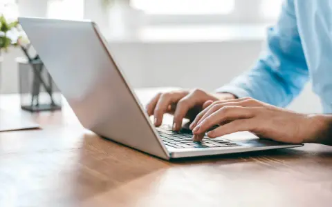 Person tippt auf einem silbernen Laptop auf einem Holztisch.