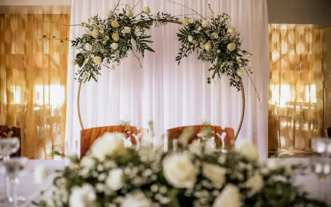 Elegancki ołtarz na ślub w plenerze, ozdobiony białymi kwiatami i zielenią na złotym okręgu.