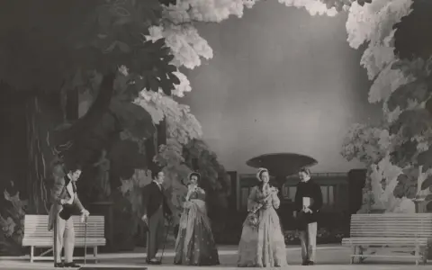 Scena z przedstawienia teatralnego, gdzie aktorzy w strojach z epoki odgrywają scenę w ogrodzie. Reżyser teatralny Szyfman zadbał o szczegóły scenografii.
