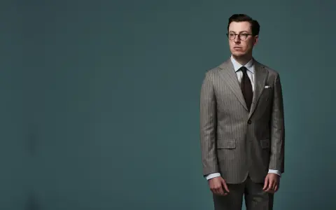 A man in a pinstripe suit, with a brown tie and glasses, stands against a teal background. The jacket length appears to be ideal, hitting just below the seat.