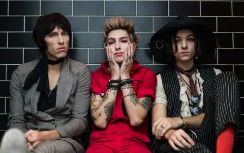 Trzech członków Palaye Royale w stylowych, rockowych strojach siedzi obok siebie na tle czarnych płytek.
